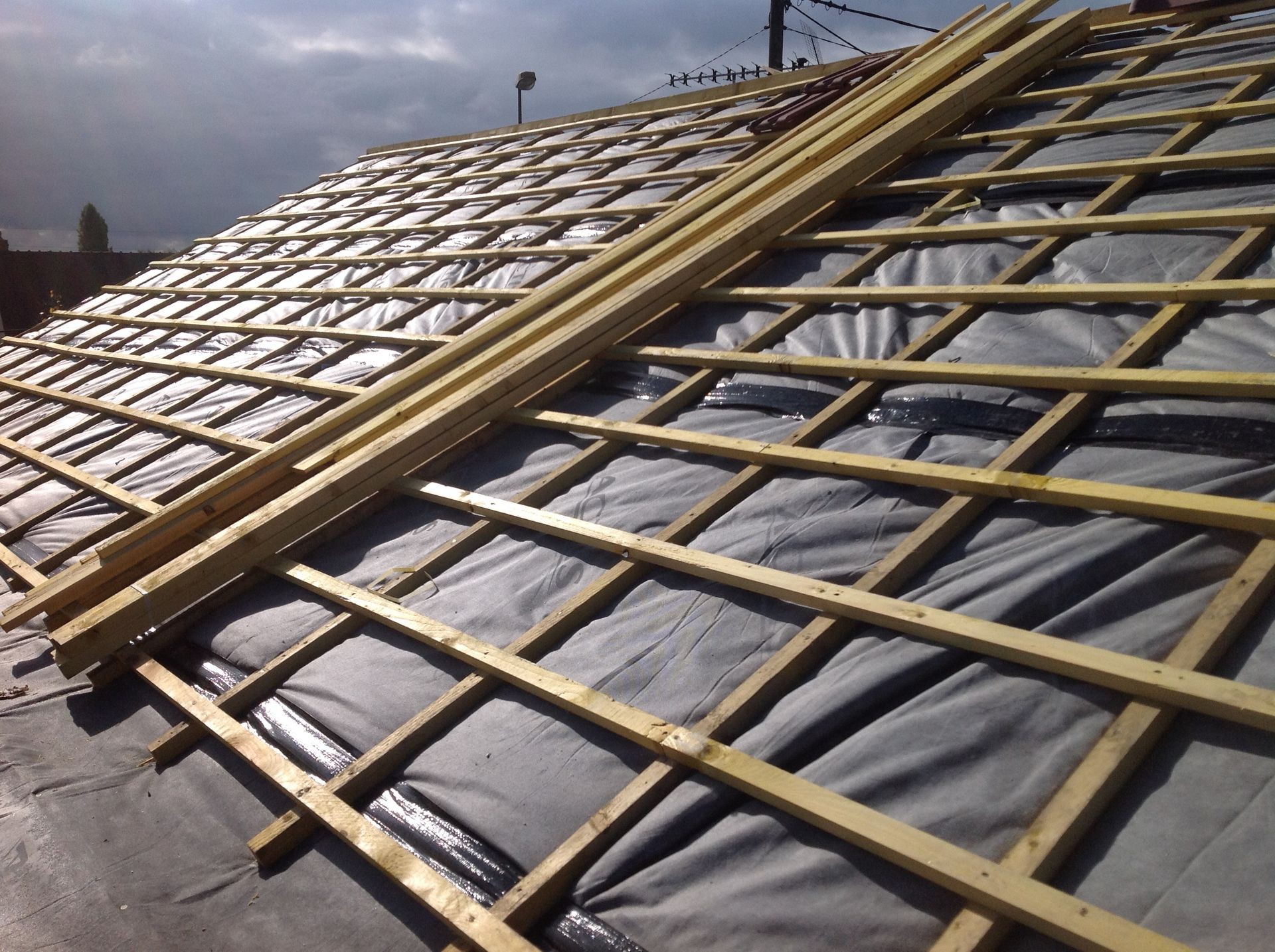 Partially constructed roof with wooden battens and black waterproof material, under a cloudy sky.